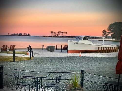 A serene waterfront scene at sunset, featuring a boat, sandy shore, and outdoor seating with a colorful sky.