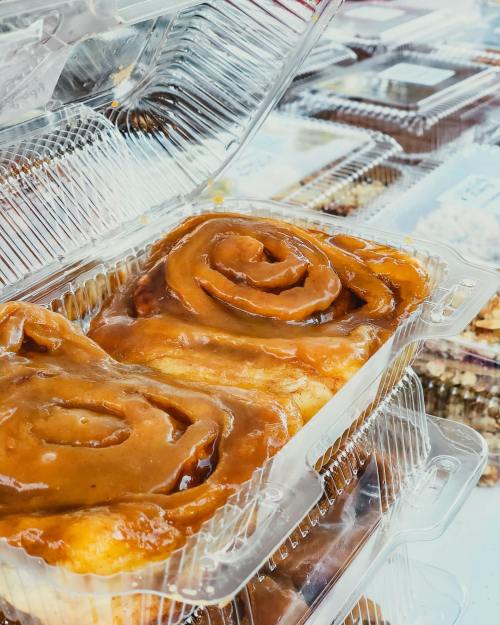 A close-up of a caramel-covered cinnamon roll in a clear plastic container, with more desserts in the background.