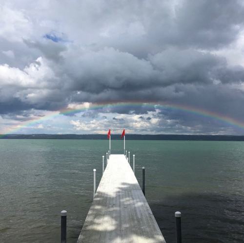 A wooden dock extends into a calm lake, with a rainbow arching over dark clouds in the background. Red flags flutter at the end.