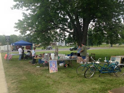 A community yard sale with tables of items, a blue tent, and people browsing, set under a large tree.
