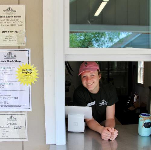 A smiling employee at a snack shack window, with a menu and hours posted nearby.