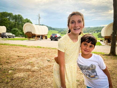 Two children smile together outdoors, with covered wagons and a scenic landscape in the background.