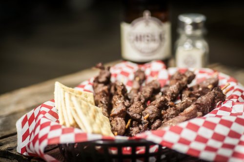 A basket of grilled meat skewers with crackers, set against a rustic background with a bottle and seasoning shaker.