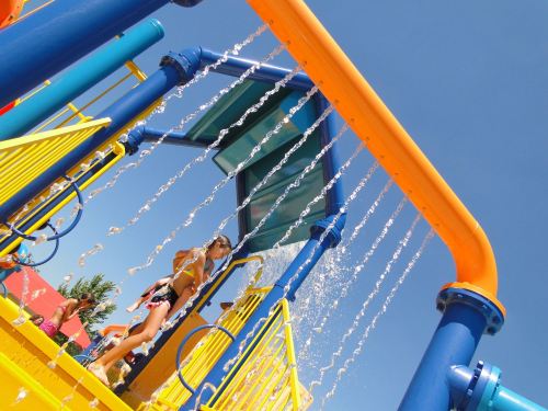 A child plays under a colorful water slide, with water cascading down in a sunny outdoor setting.