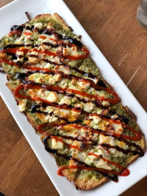 A flatbread topped with pesto, cheese, and drizzled with sauces on a white plate, set on a wooden table.