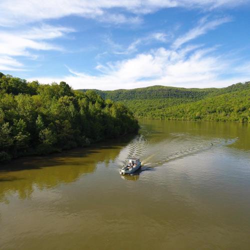 A boat glides across a calm river surrounded by lush green hills and a clear blue sky.