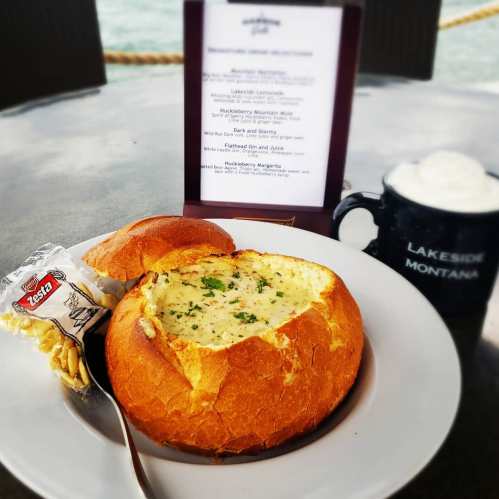 A bread bowl filled with creamy soup, accompanied by a roll, a packet of crackers, and a mug of lemonade.