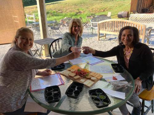 Three women toast with drinks at a table, surrounded by a cheese platter and outdoor seating in a scenic setting.