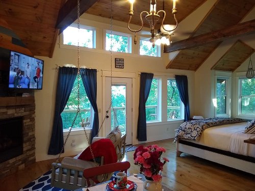 Cozy cabin interior with a swing chair, fireplace, large windows, and a bed, featuring a TV and a dining table with flowers.