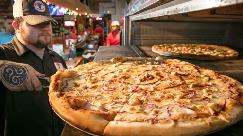 A chef pulls a large pizza from an oven, with another pizza visible behind it in a bustling pizzeria.