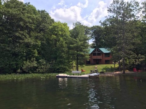 A serene lakeside view featuring a cabin, lush trees, and a small boat docked near the shore.