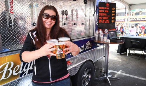 A woman in sunglasses holds three beers in front of a beverage truck at an outdoor event.