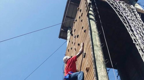 A person in a red shirt climbs a tall wooden climbing wall on a sunny day.
