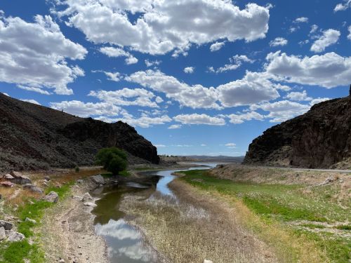 A serene landscape featuring a winding river, grassy banks, and rocky hills under a blue sky with fluffy clouds.