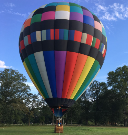A colorful hot air balloon stands on the ground against a blue sky and green trees.