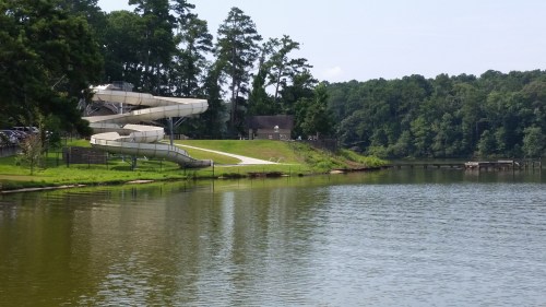A calm lake with a water slide in the background, surrounded by trees and a grassy area.