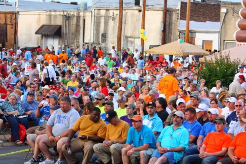A large crowd of people in colorful shirts gathered outdoors for an event, with tents and buildings in the background.