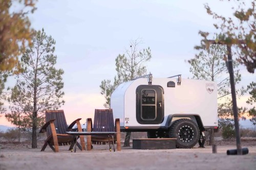 A compact trailer parked in a serene outdoor setting with wooden chairs and a fire pit, surrounded by trees at sunset.