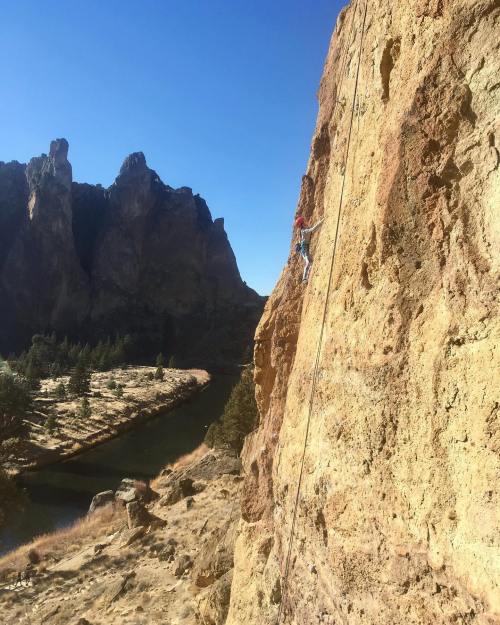 A climber ascends a rocky cliff with mountains and a river in the background under a clear blue sky.