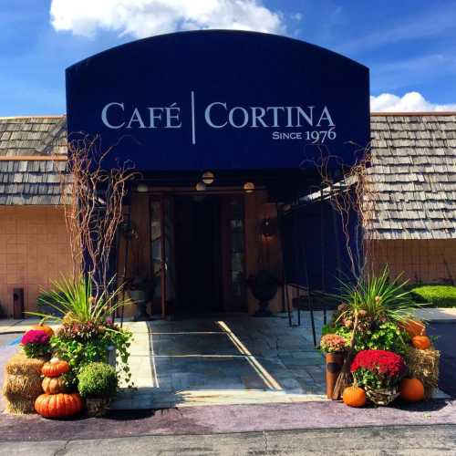 Entrance of Café Cortina with a blue awning, pumpkins, and colorful flowers, set against a clear sky.