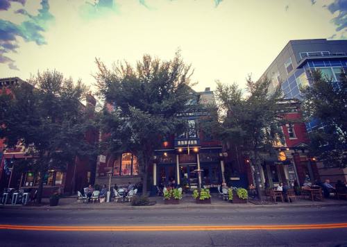 A charming restaurant with outdoor seating, surrounded by trees and colorful planters, on a lively street at dusk.