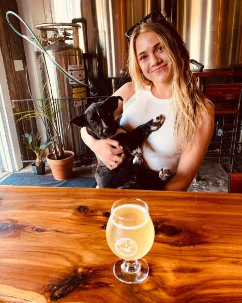 A woman with long hair holds a black puppy while sitting at a table with a glass of beer in front of her.