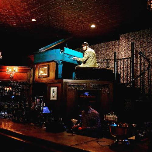 A man plays a blue piano on a raised platform in a dimly lit bar, with a bartender mixing drinks below.