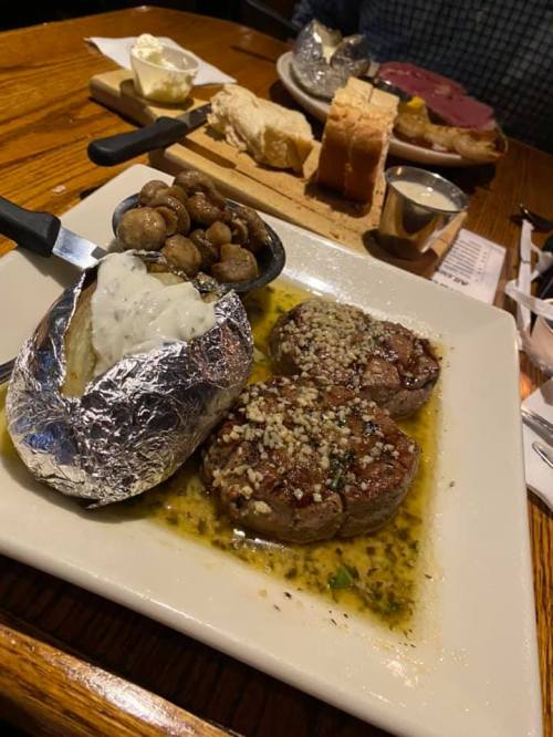A plate featuring two filet mignon steaks, a baked potato with sour cream, sautéed mushrooms, and bread in the background.