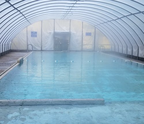 Indoor swimming pool under a clear, arched plastic roof, with calm water and a misty atmosphere.