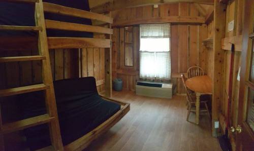 Cozy wooden cabin interior with bunk beds, a futon, a table, and a window with checkered curtains.