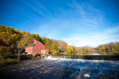 A red mill beside a flowing river, surrounded by colorful autumn trees under a clear blue sky.