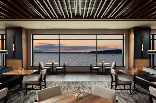 Modern restaurant interior with wooden tables, chairs, and large windows showcasing a sunset over the water.