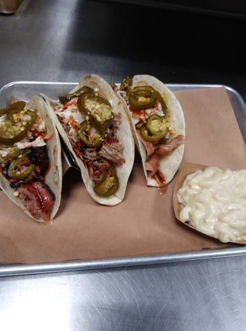 Three tacos filled with meat, topped with jalapeños, served with a side of creamy macaroni salad on a tray.