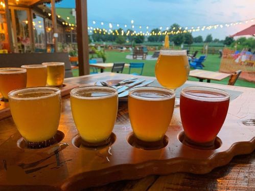 A wooden flight of craft beers in various colors, set on a table with a scenic outdoor background and string lights.