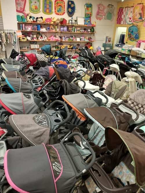 A cluttered store aisle filled with various strollers and baby gear, with shelves of toys in the background.