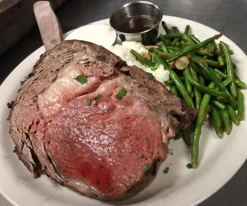 A plate featuring a large slice of medium-rare prime rib, green beans, and a small cup of sauce.