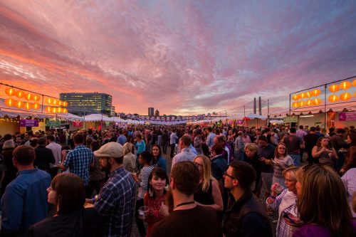 A vibrant outdoor festival scene at sunset, with a large crowd enjoying food and drinks under colorful tents.