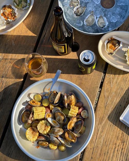 A bowl of seafood with corn and potatoes, surrounded by drinks and oysters on a wooden table.