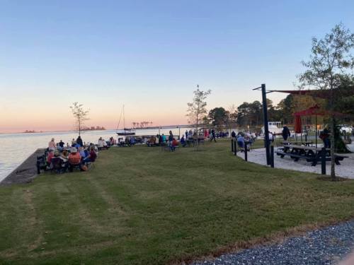 A waterfront scene with people gathered on the grass, enjoying the sunset near a calm lake. Trees and picnic tables are visible.