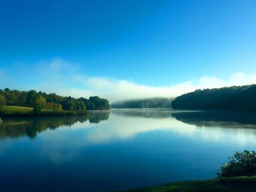 A serene lake reflects a clear blue sky and misty hills, surrounded by lush greenery.
