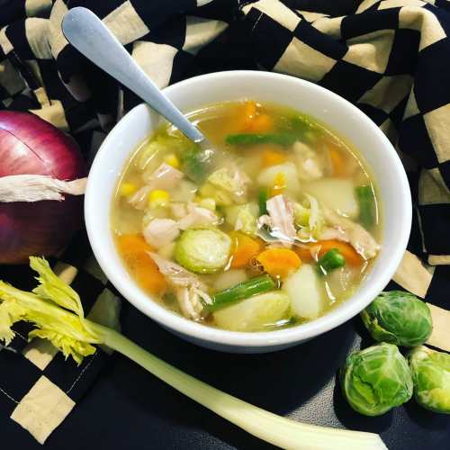 A bowl of hearty chicken soup with vegetables, including carrots, celery, and Brussels sprouts, on a checkered cloth.