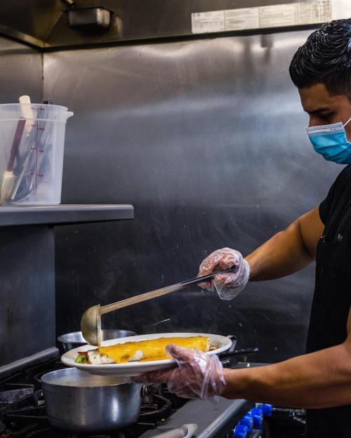 A chef in a mask and gloves serves a dish from a pot in a busy kitchen.
