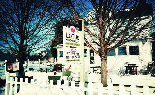 A snowy scene featuring a sign for Lotus Kitchen, LLC, with a white fence and trees in the background.