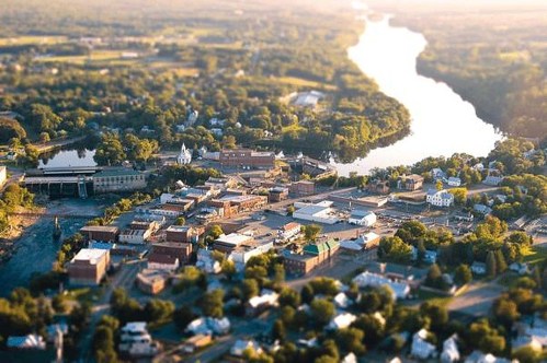 Aerial view of a small town by a river, featuring buildings, trees, and a scenic landscape in warm sunlight.