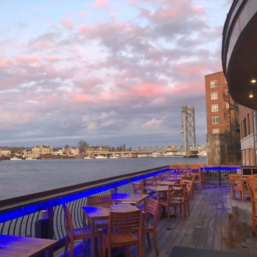 A waterfront view with wooden tables, a bridge in the background, and colorful clouds at sunset.