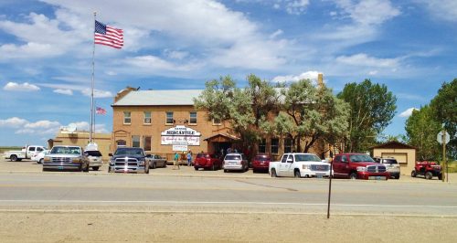 A historic building with a sign reading "Mercantile," surrounded by parked cars and a flag flying in the background.