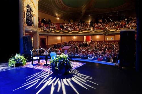 A stage set with chairs and plants, facing a packed theater audience in a beautifully decorated venue.