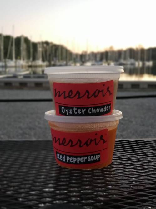 Two soup containers labeled "Oyster Chowder" and "Red Pepper Soup" sit on a table with a marina in the background.