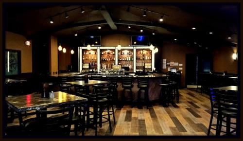 A dimly lit bar interior featuring a wooden floor, high tables, and a well-stocked backlit bar.