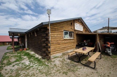 A rustic wooden building with a sign, picnic table, and parked motorcycles under a cloudy sky.